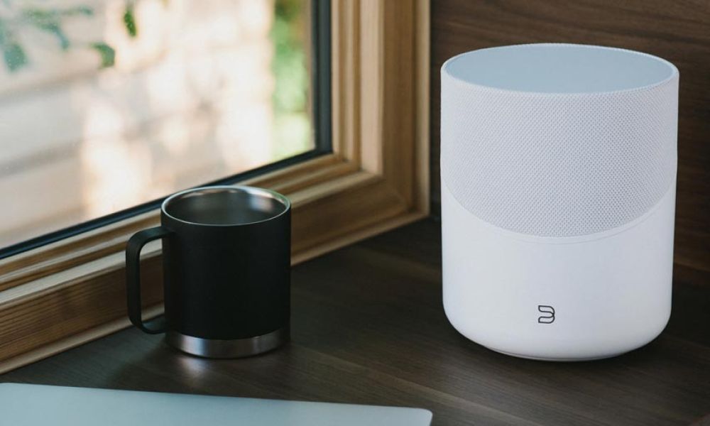 White wireless speaker next to a black mug on a wooden desk by a window