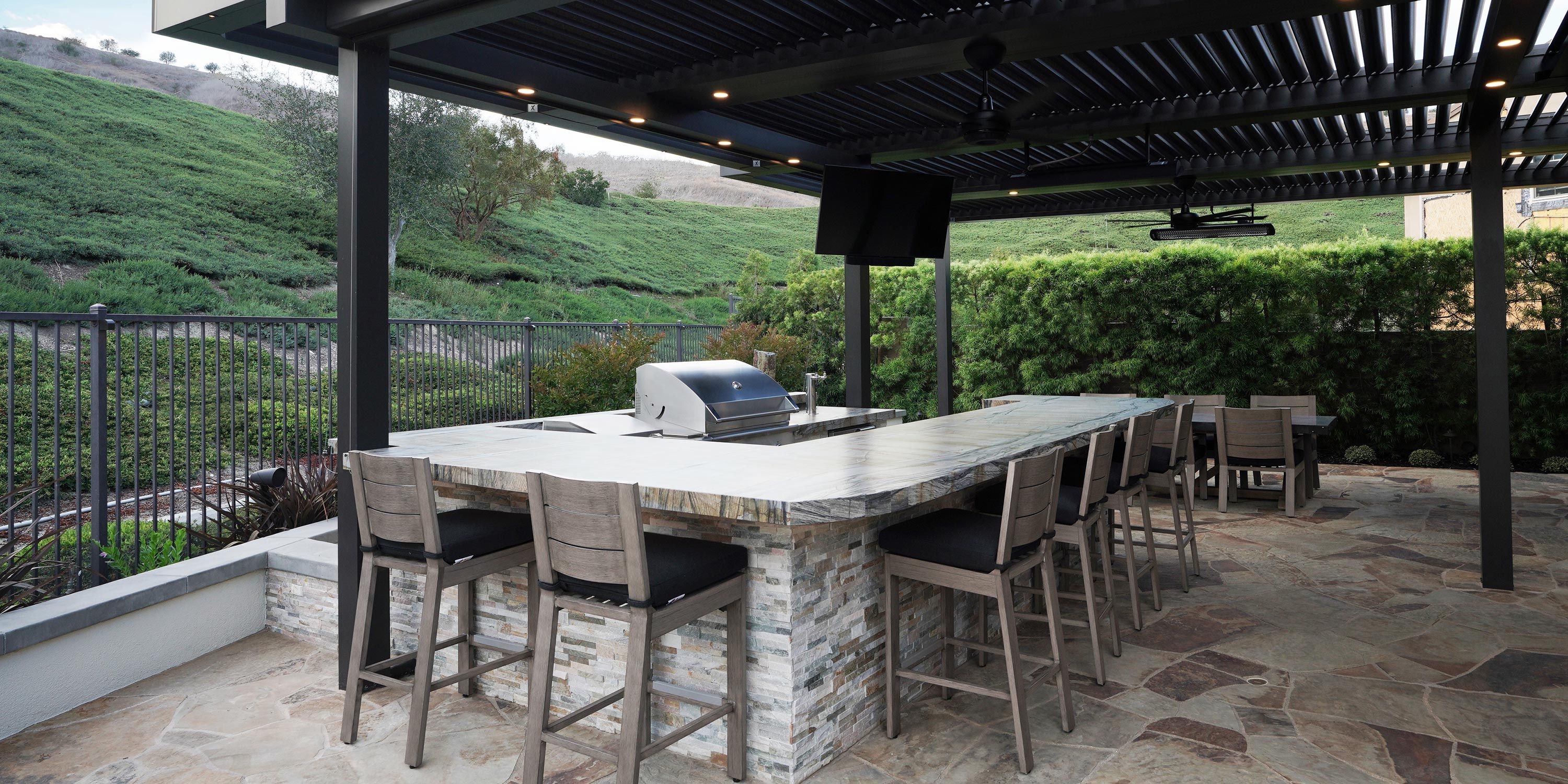 An outdoor kitchen with a barbecue grill and bar seating, surrounded by lush greenery and a pergola overhead.