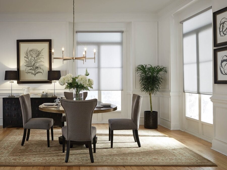 A dining room space with a dining table and chairs, lamps on a cabinet in the background, and two windows featuring motorized window treatments.
