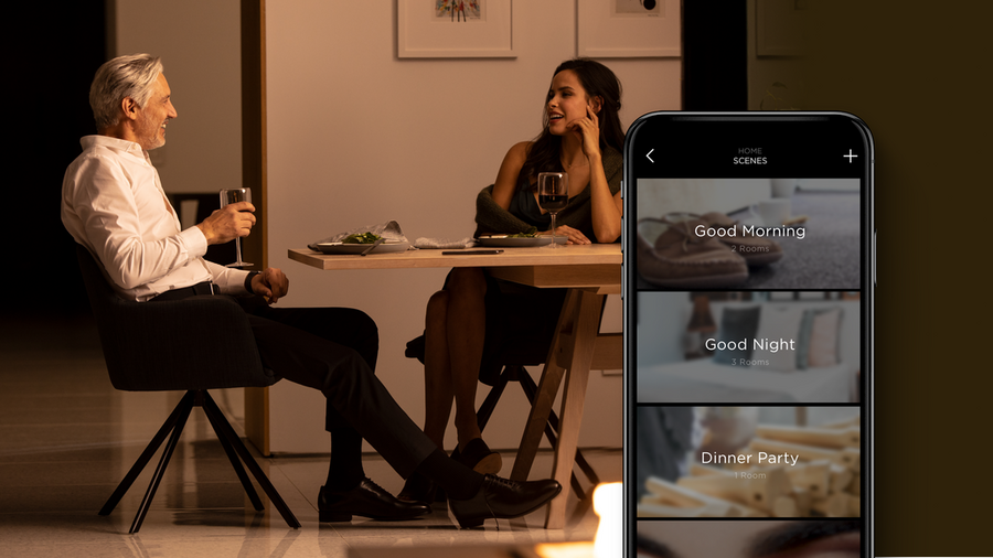 A couple sitting at a table with a zoomed-in smartphone in the foreground showing a Savant smart home control interface.