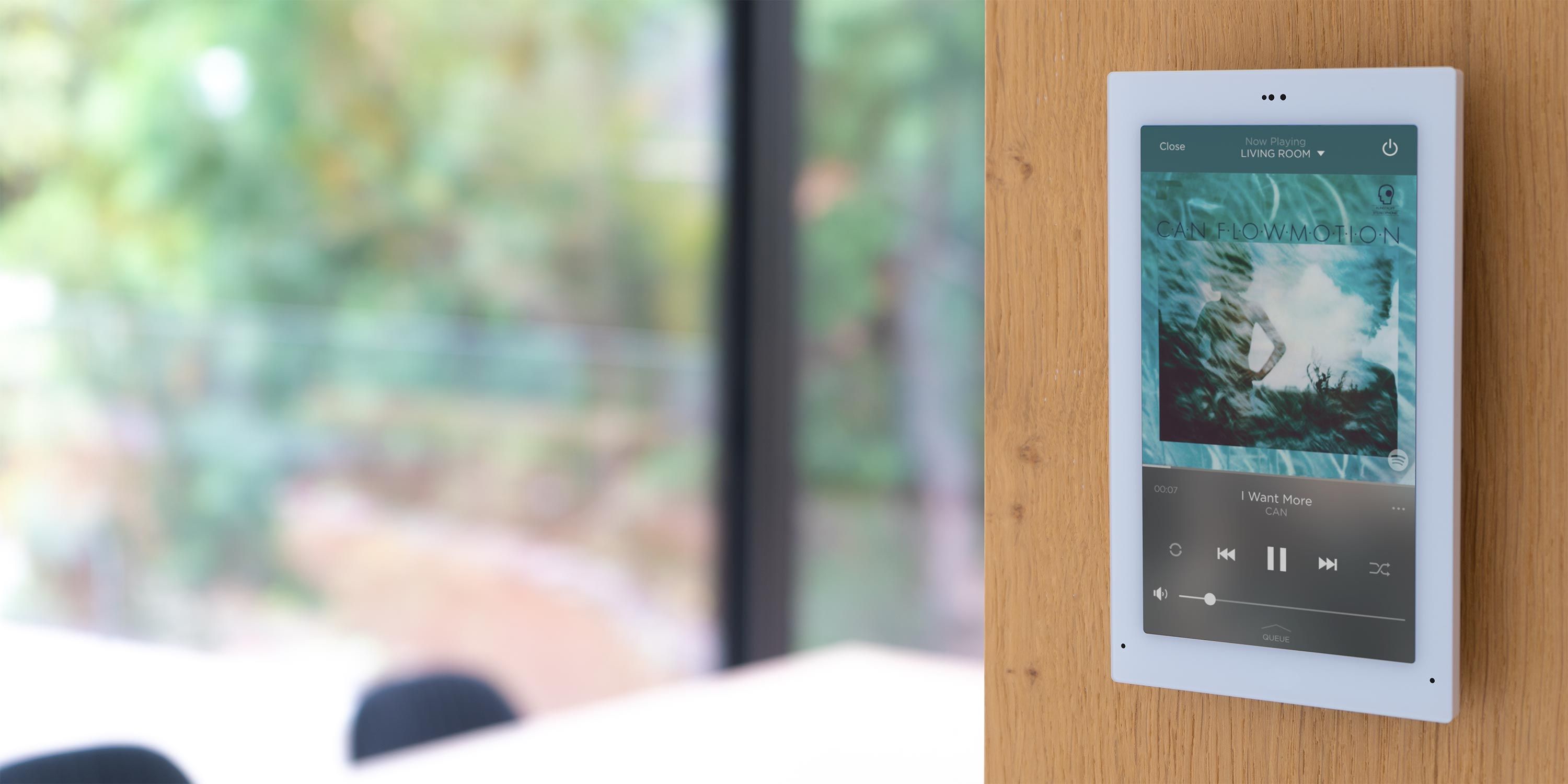 Wall-mounted touchscreen displaying music controls and album art in a bright, modern home.