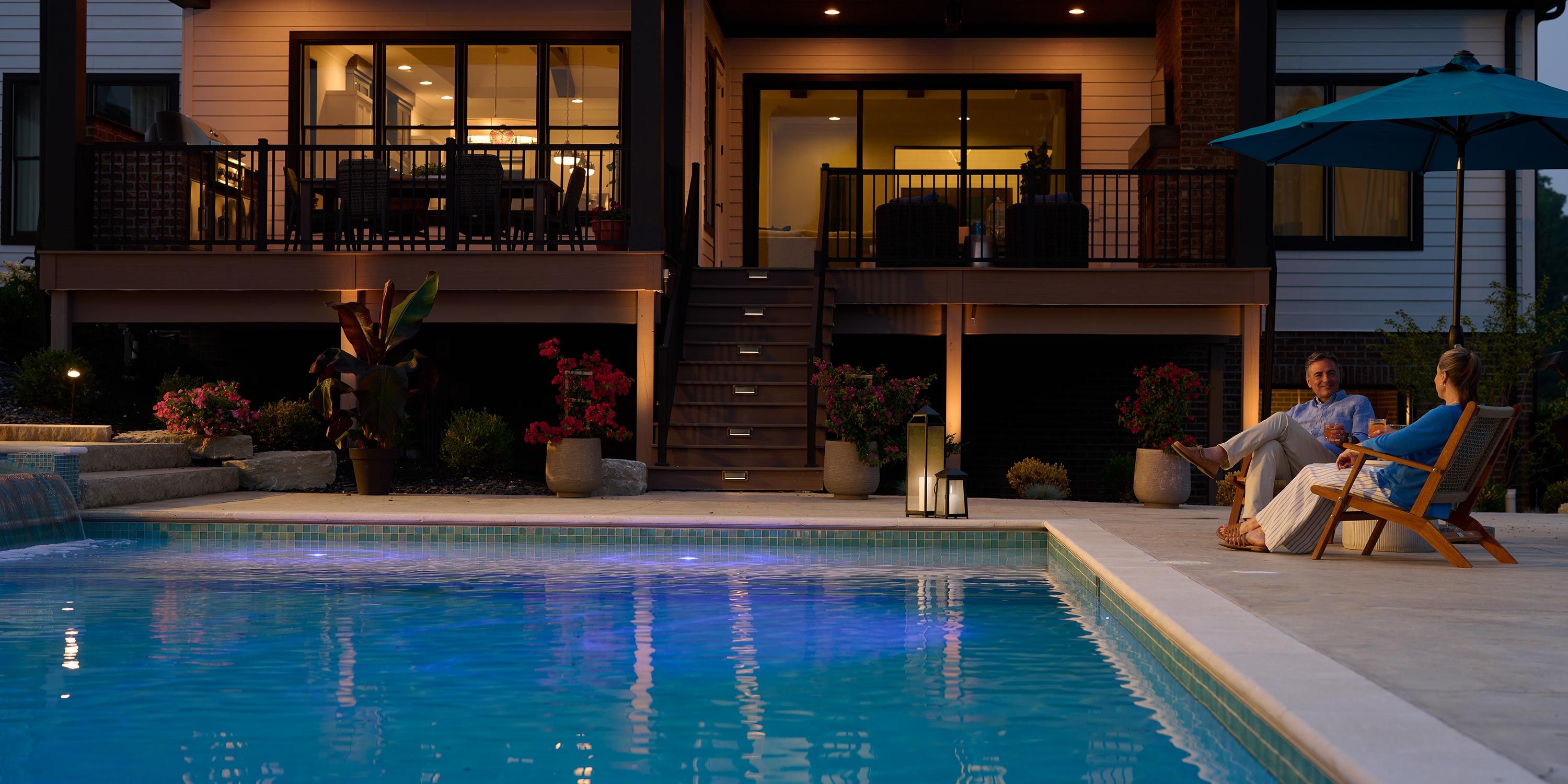 Evening view of a backyard pool area with a seating area, illuminated pool, and a couple relaxing by the poolside.