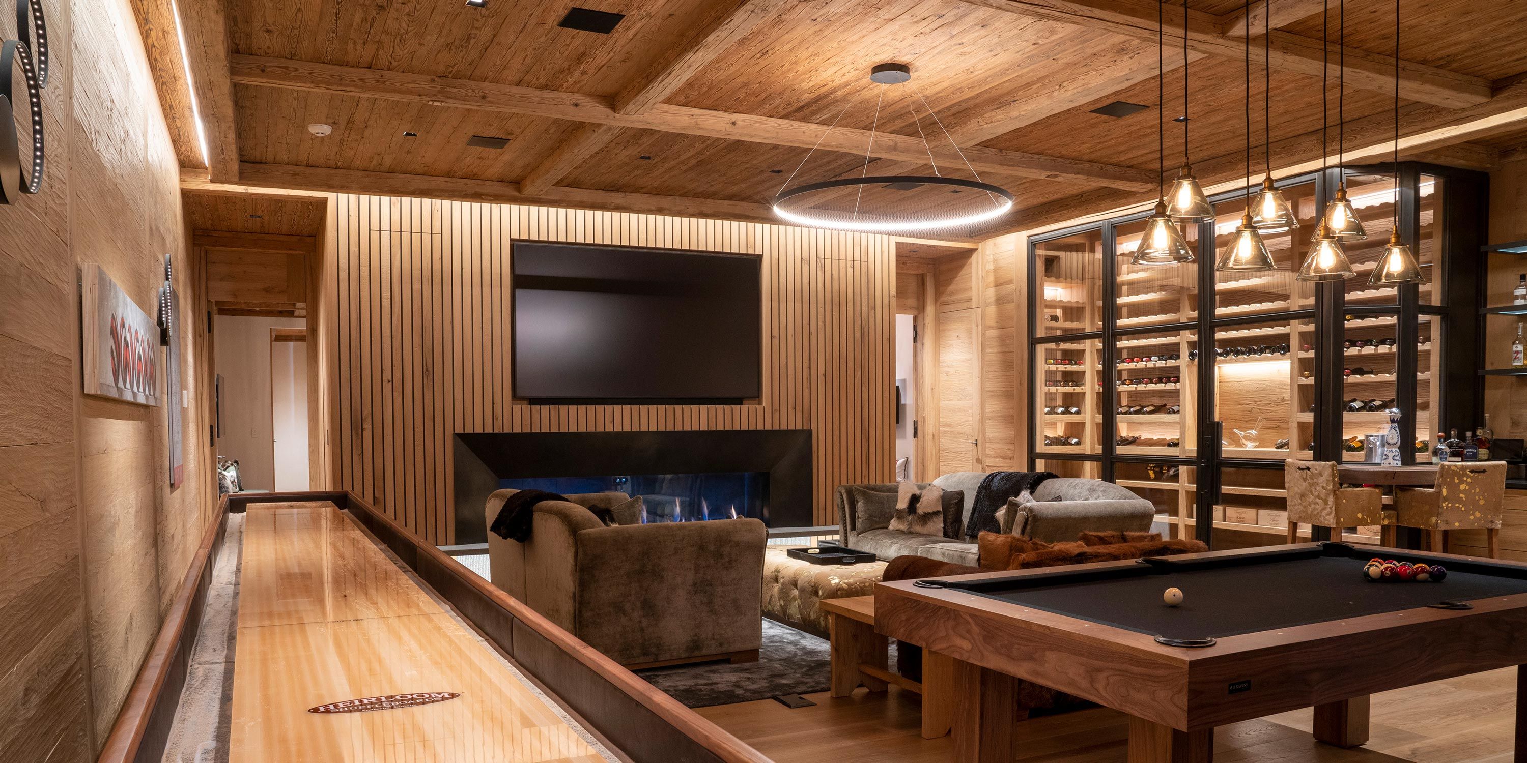 Cozy game room with a wooden shuffleboard table, pool table, large TV, fireplace, and a wine rack, illuminated by warm pendant lights.