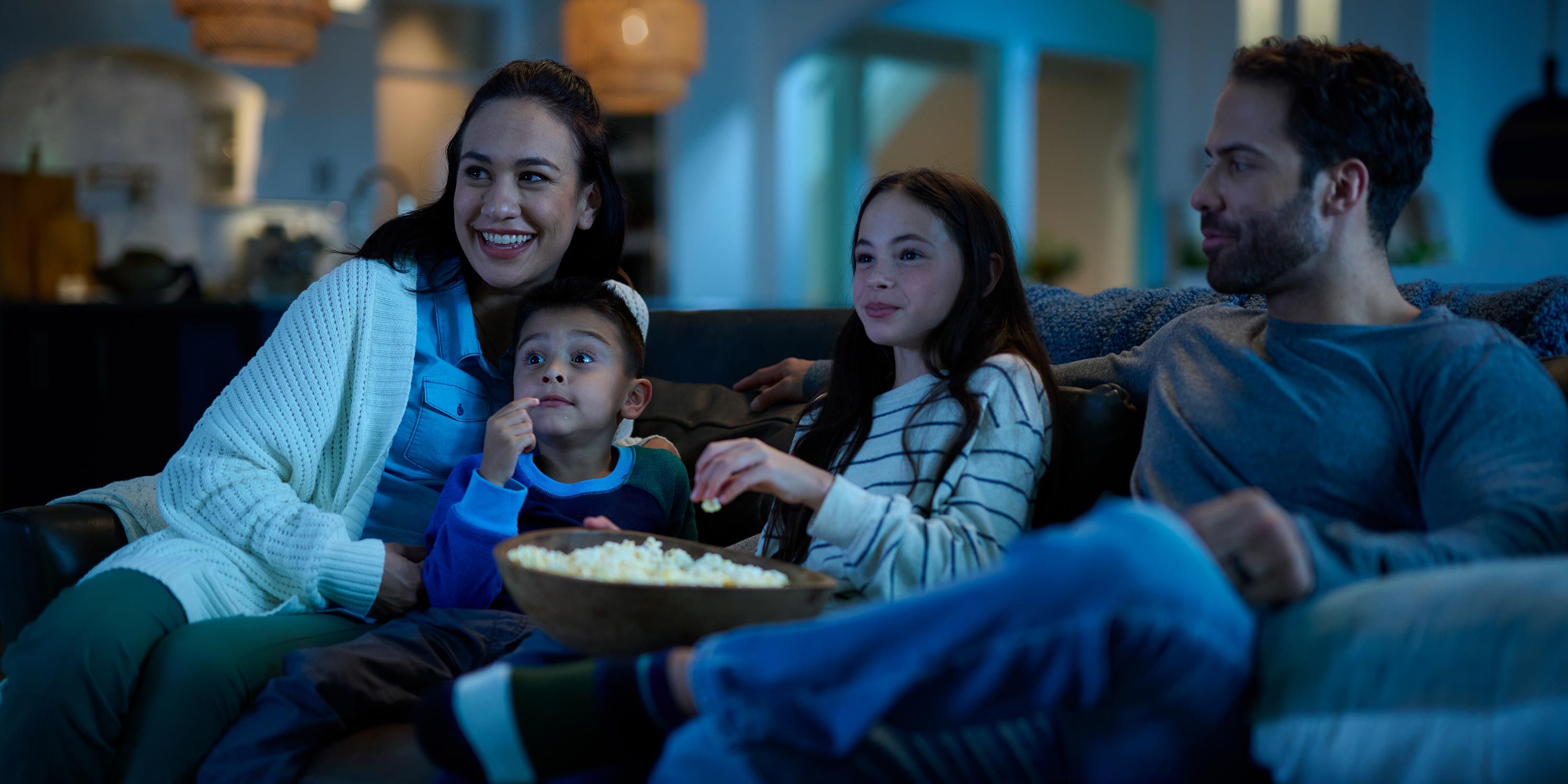 Family enjoying a movie night on the couch with a bowl of popcorn, illuminated by the soft glow of the television.